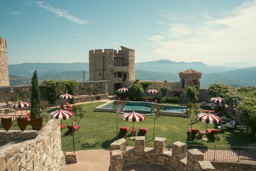 Steinschloss mit Panoramaschwimmbad, gepflegtem Garten und roten Sonnenschirmen, eingebettet in die Berge und umgeben von einer atemberaubenden Landschaft.