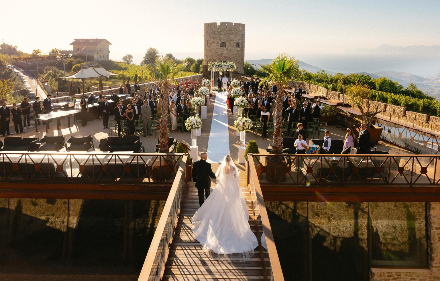 Sposi percorrono la passerella verso una cerimonia nuziale all’aperto in un castello panoramico, circondati dagli invitati e da eleganti allestimenti floreali.