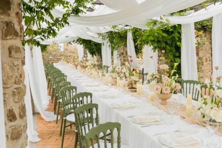 Tavolata nuziale all’aperto in un’elegante location in pietra, con sedie verde salvia, fiori freschi e drappeggi bianchi sotto il pergolato.