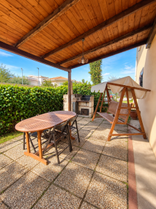 Grande terrasse extérieure aménagée avec une table et des chaises en bois, une balançoire couverte et un barbecue maçonné.