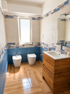 Salle de bains moderne avec carrelage beige et bleu, meuble vasque en bois, sanitaires blancs et sol imitation bois.