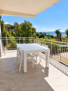 Terrasse panoramique avec vue sur la mer, table et chaises blanches, entourée de verdure.
